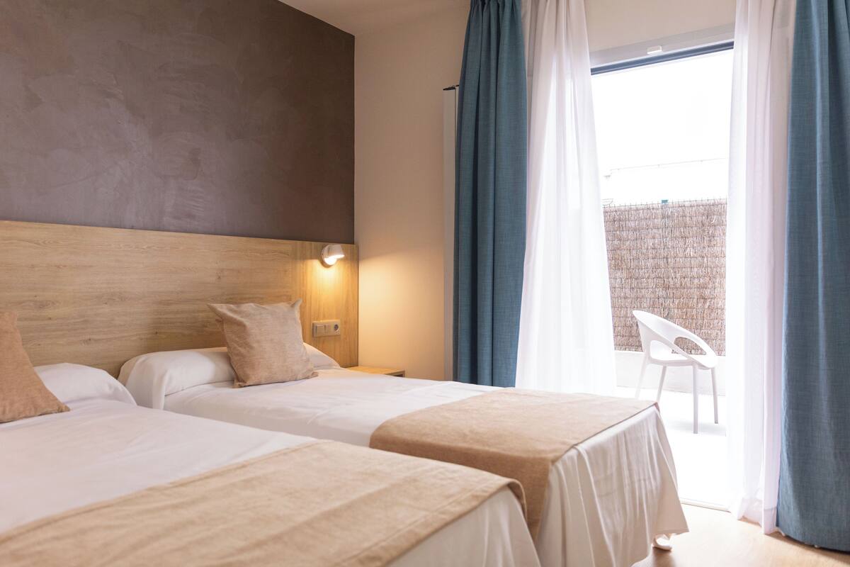 A hotel room with two beds, a wooden headboard, a nightstand with a lamp, and a view of a balcony with a chair.
