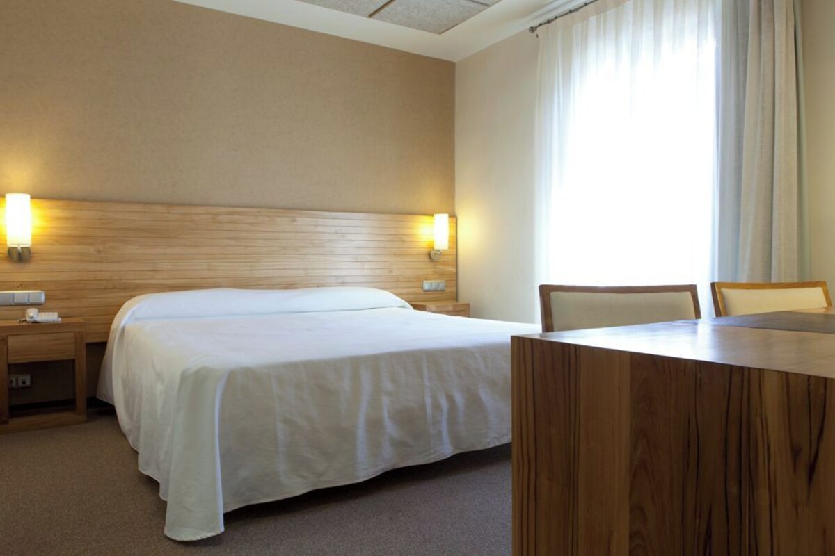A hotel room with a large bed, a wooden headboard, two bedside lamps, a wooden desk, and a chair.