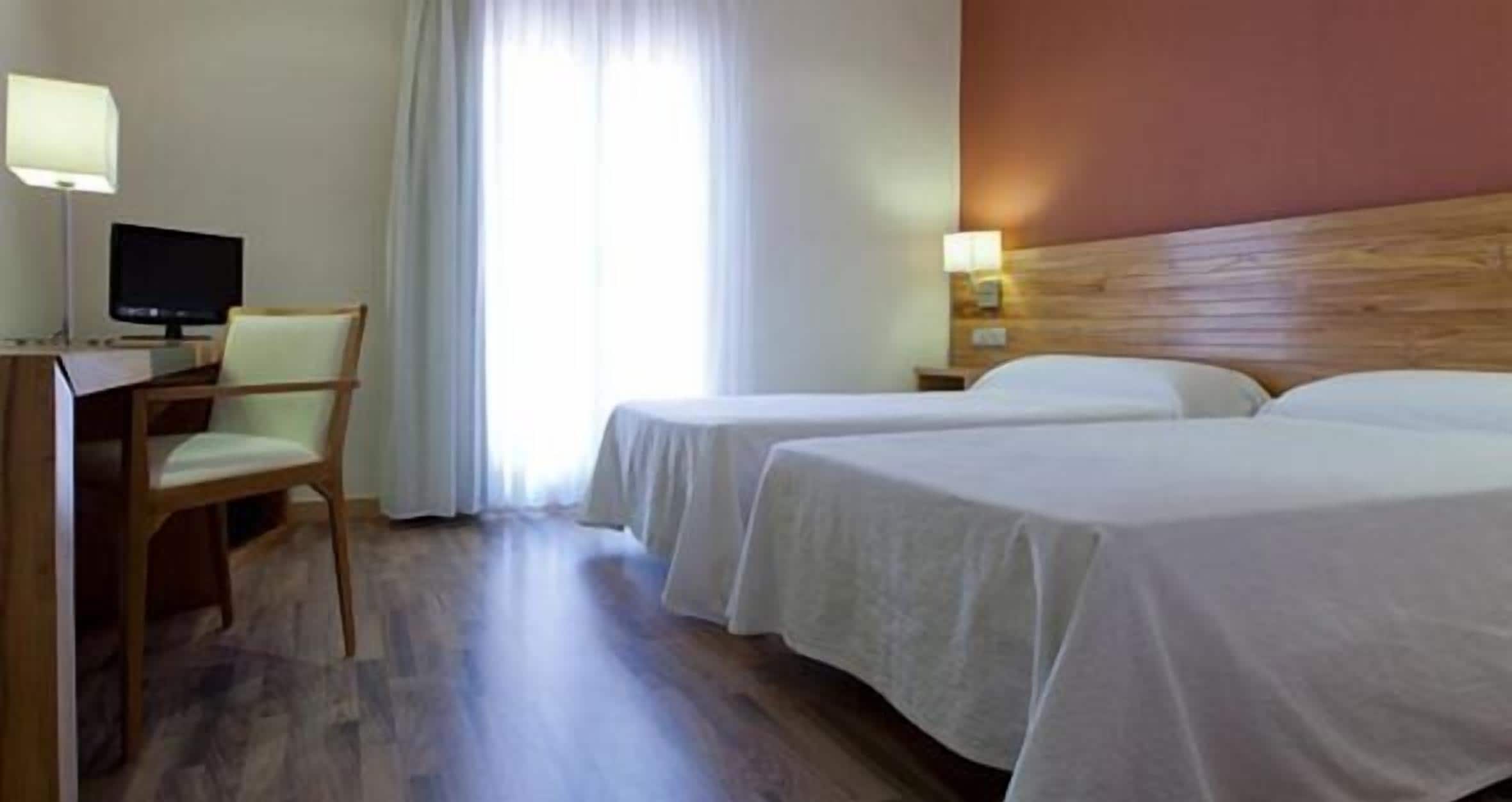 A hotel room with two beds, a desk with a computer, a chair, and a window with curtains.