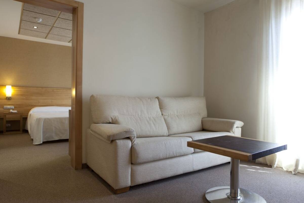 A hotel room with a beige sofa, a small table, a bed, and a nightstand.