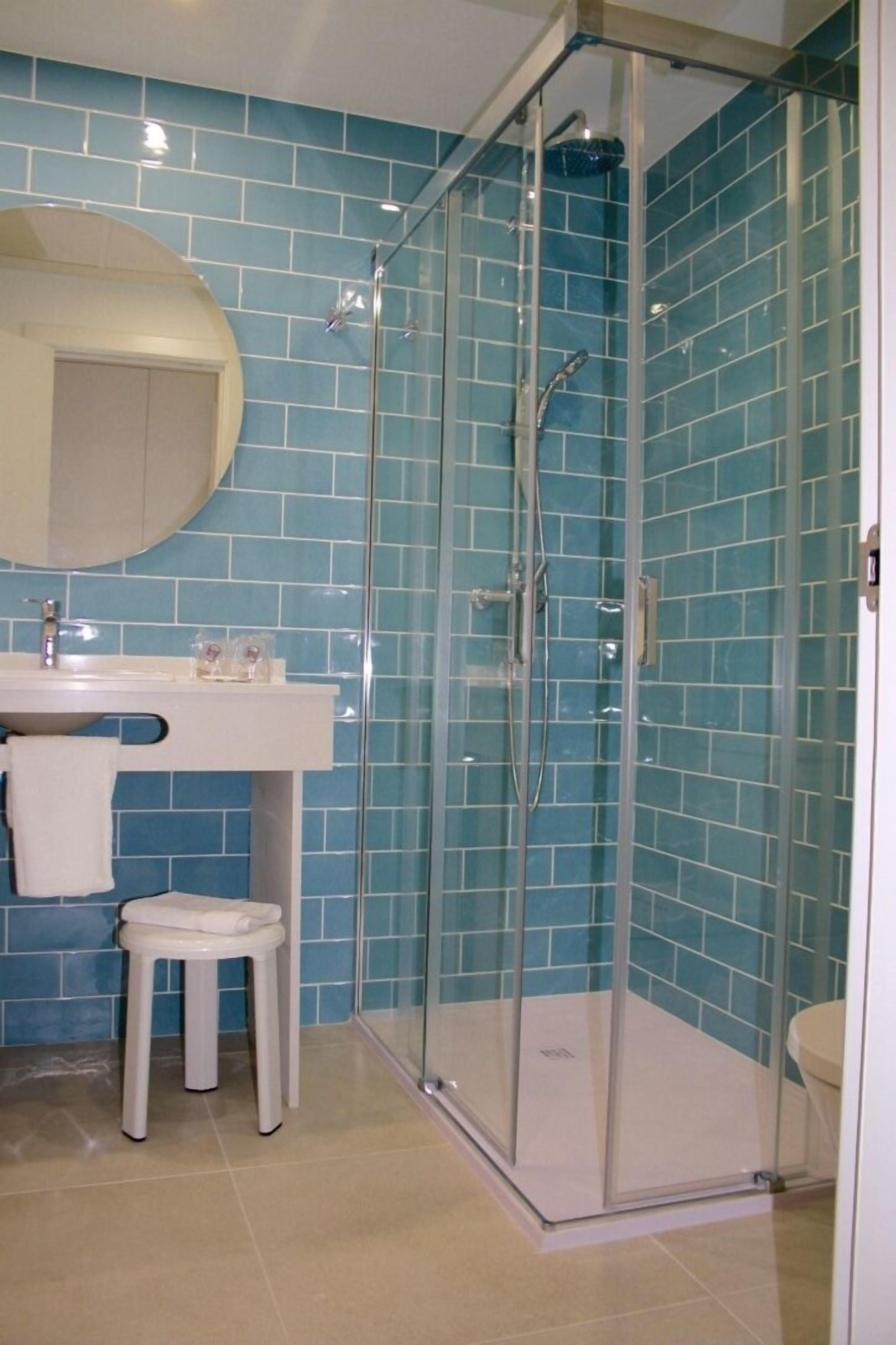 A bathroom with a glass-enclosed shower, a sink with a mirror, and a stool.
