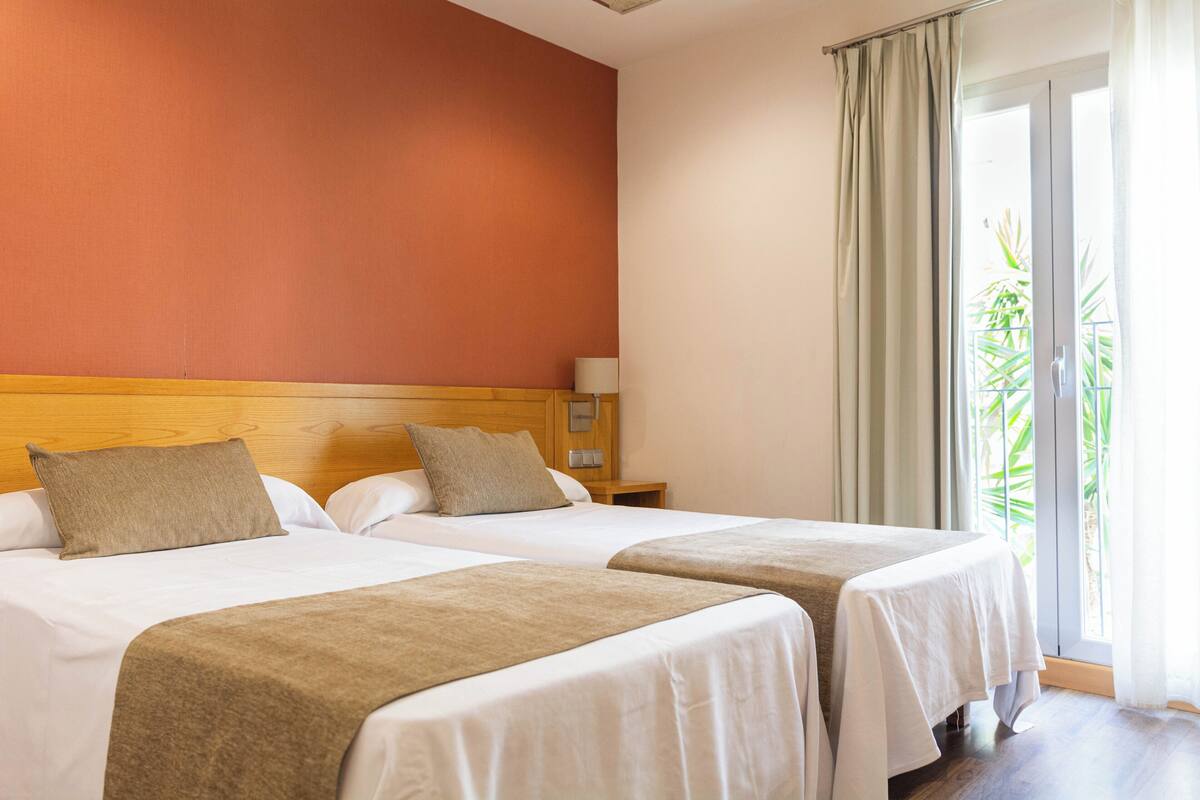 A hotel room with two beds, a wooden headboard, a nightstand with a lamp, and a view of greenery through a sliding door.