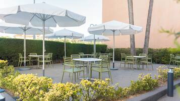 An outdoor patio area with multiple tables and chairs, surrounded by white umbrellas and greenery.