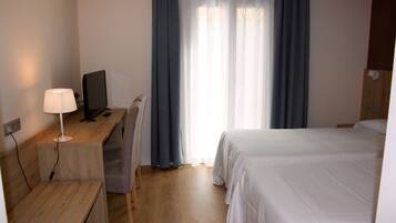 A hotel room with a wooden desk, a computer, a chair, a bed, and a window with curtains.