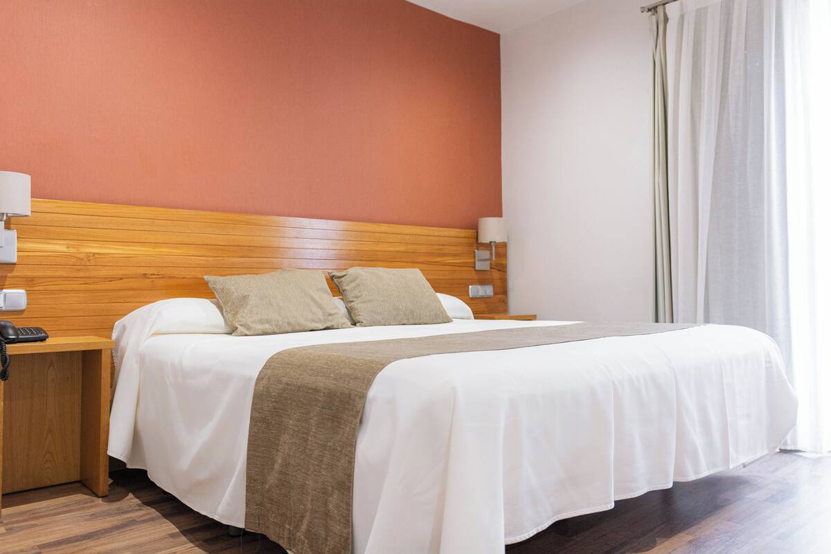 A hotel room with a wooden headboard, white bedding, a brown throw blanket, and a nightstand with a phone.