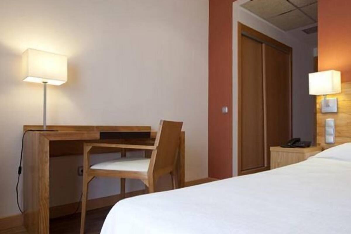 A hotel room with a bed, a desk with a chair, and two bedside lamps.