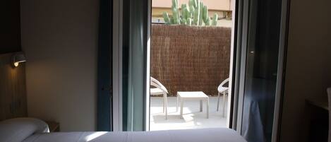 Habitación de hotel con cama, mesita de noche, escritorio y silla. Posee una puerta corrediza de vidrio que da a un espacio exterior con un cactus y una mesa.