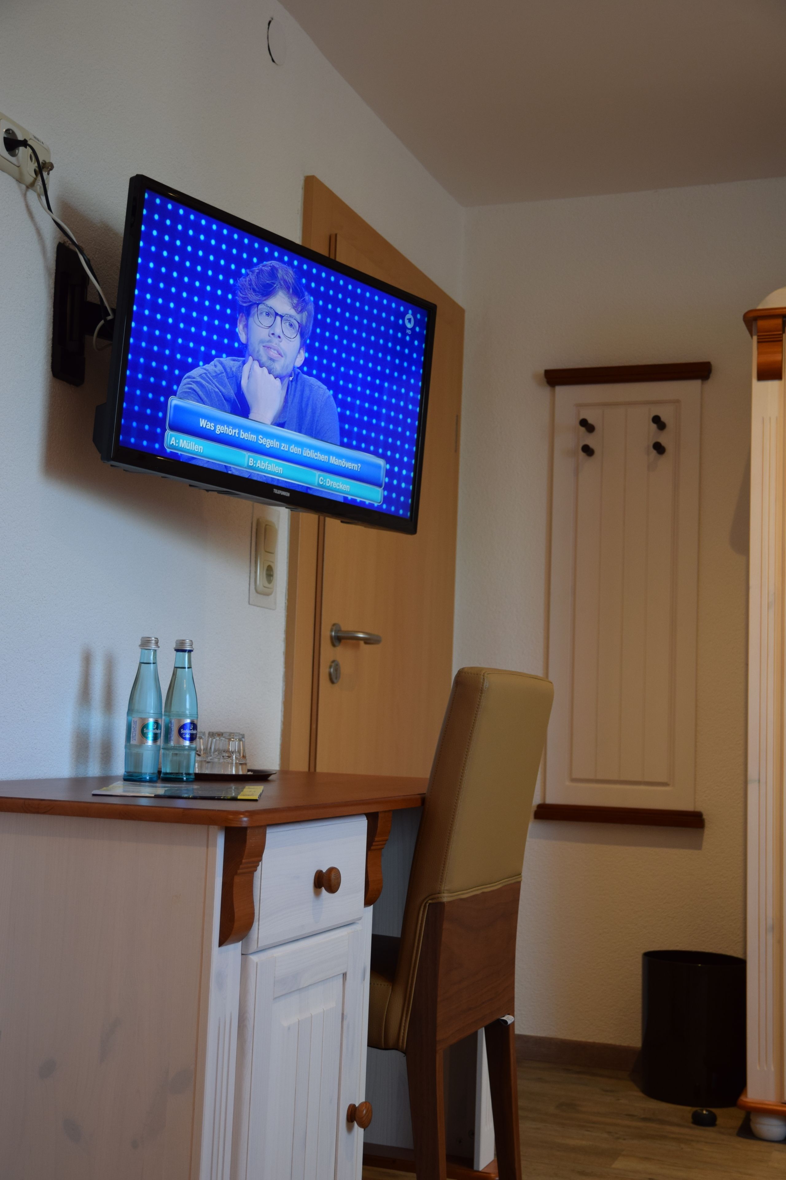 A room with a television mounted on the wall displaying a quiz. There is a wooden desk with a chair, a cabinet, and two bottles on the counter. A trash can is visible in the corner.