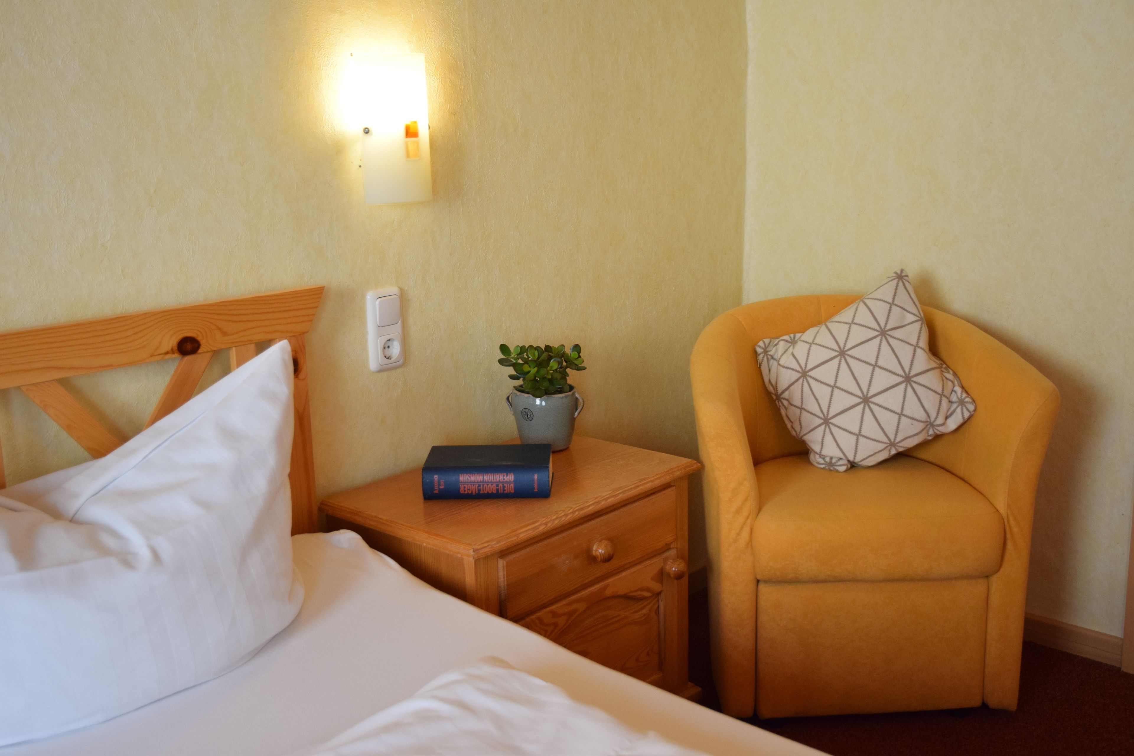 Une chambre d’hôtel avec un lit, une table de chevet sur laquelle se trouvent un livre et une plante, et une chaise agrémentée d’un coussin aux motifs géométriques.
