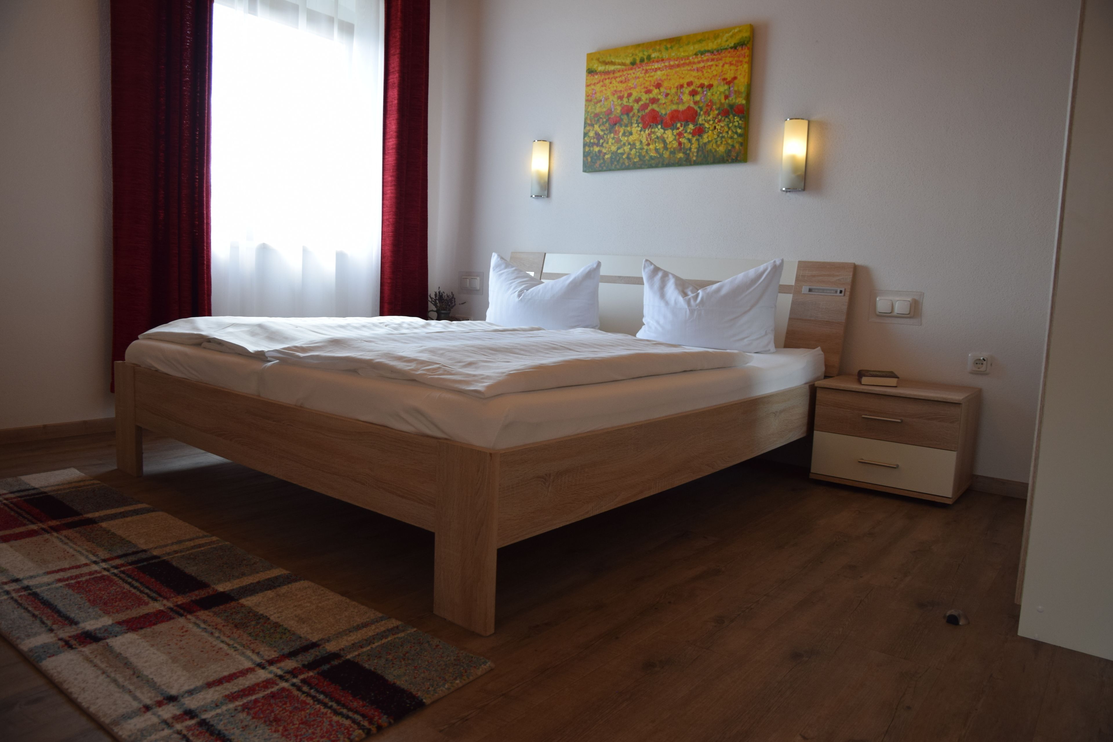A modern hotel room with a wooden bed, a plaid area rug, a nightstand, a wall-mounted lamp, and a painting.