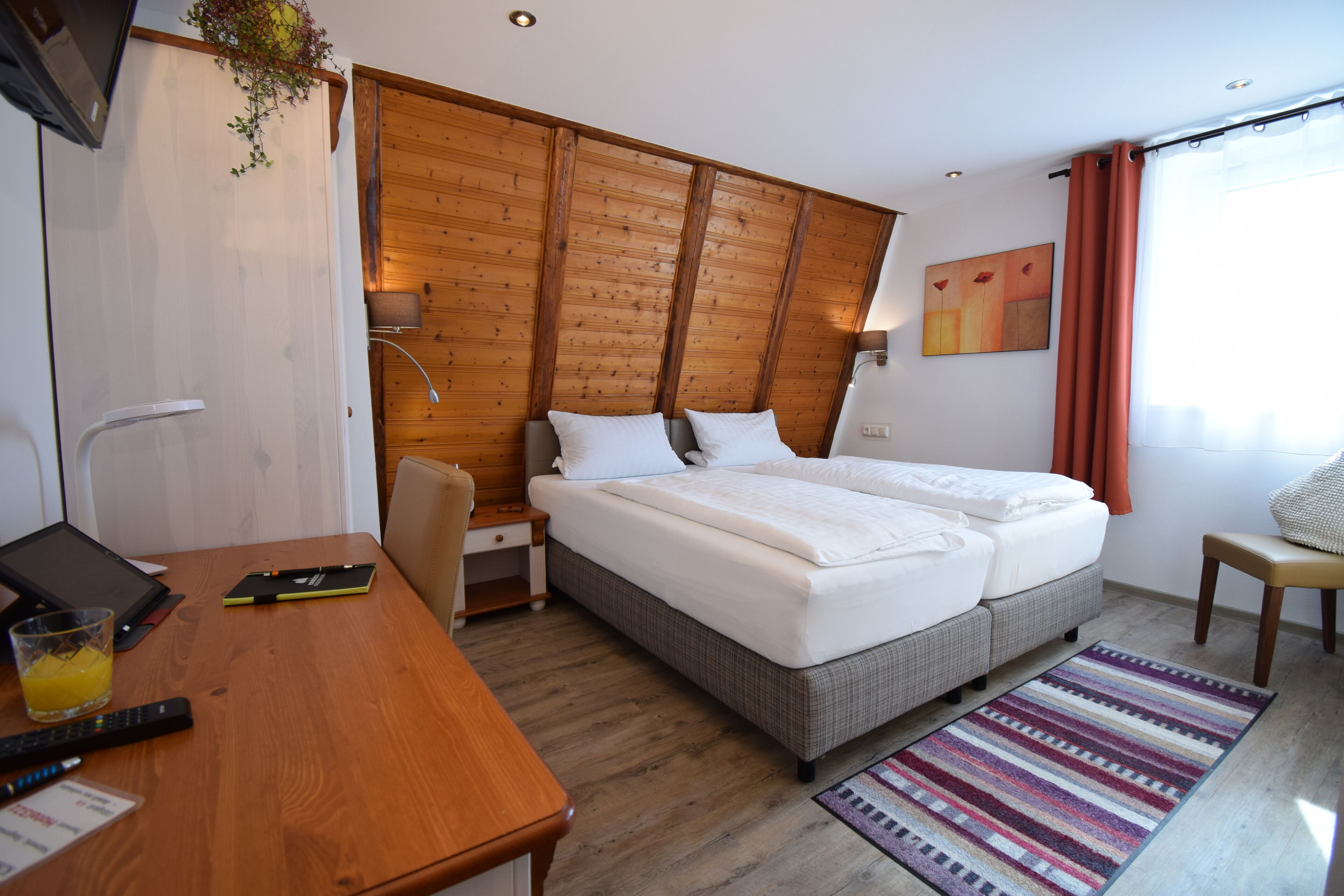 A hotel room with a wooden headboard, a bed with white linens, a desk with a laptop, and a colorful rug.