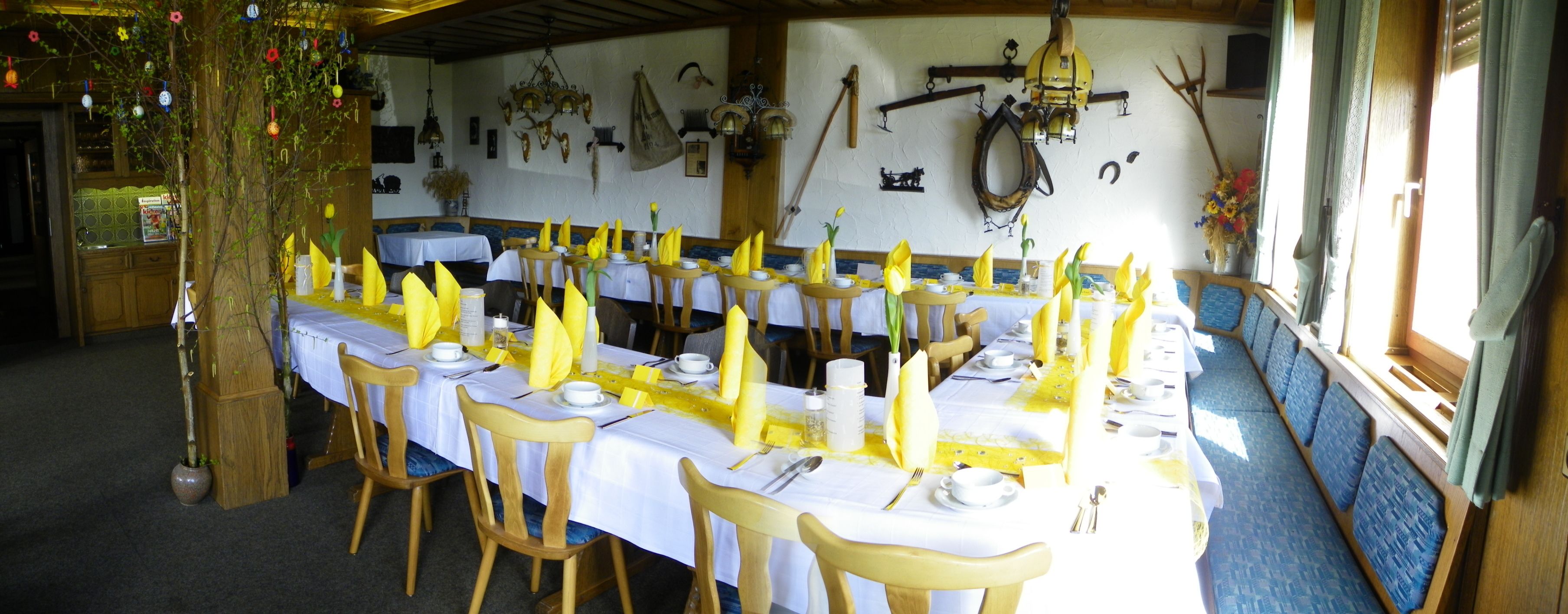 Uma longa mesa de jantar posta com guardanapos amarelos, toalhas de mesa brancas e cadeiras de madeira em um ambiente rústico com teto e paredes decorados com diversos enfeites.