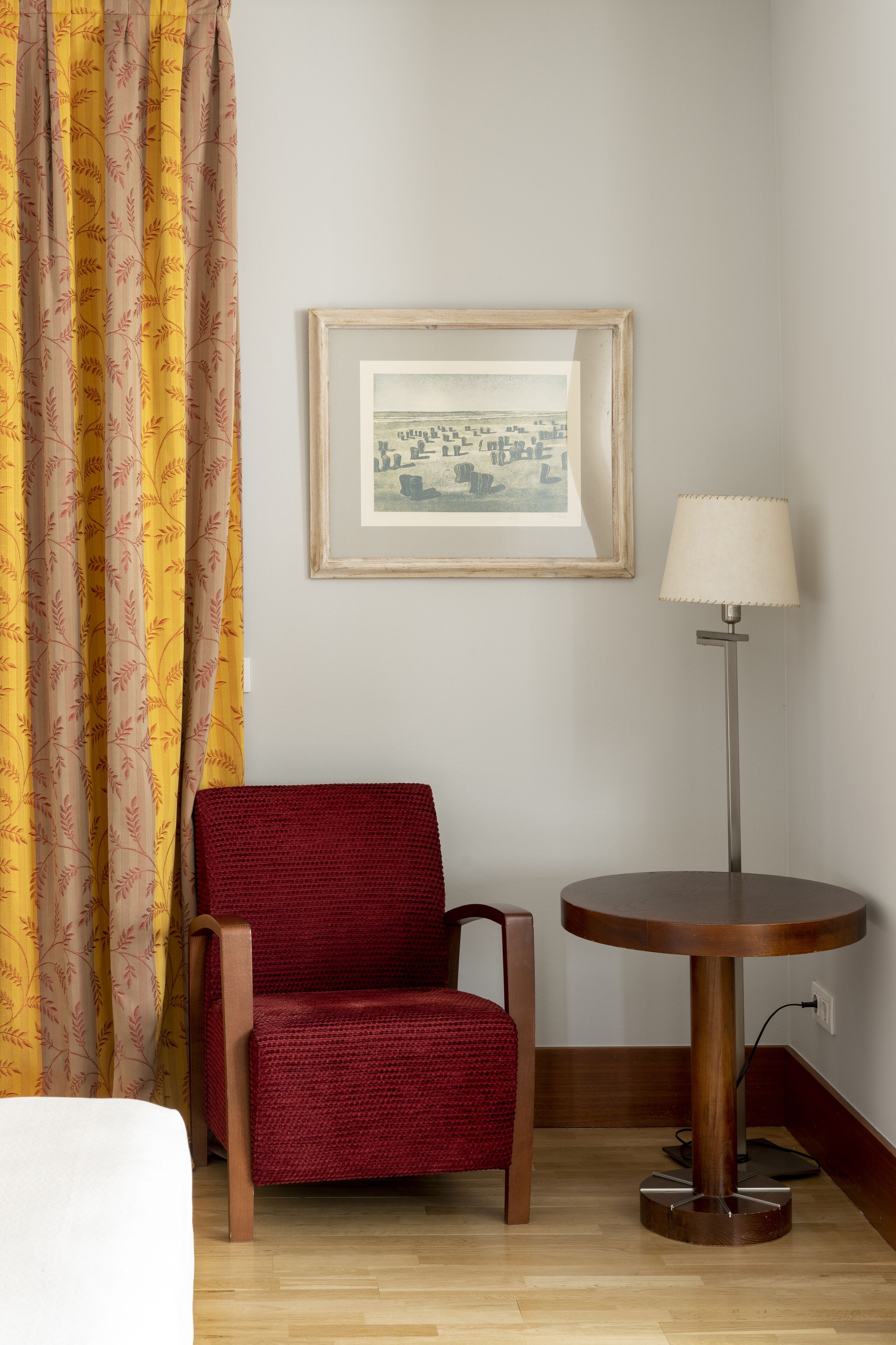 A room with a wooden chair, a round table with a lamp, a framed picture on the wall, and a window with curtains.