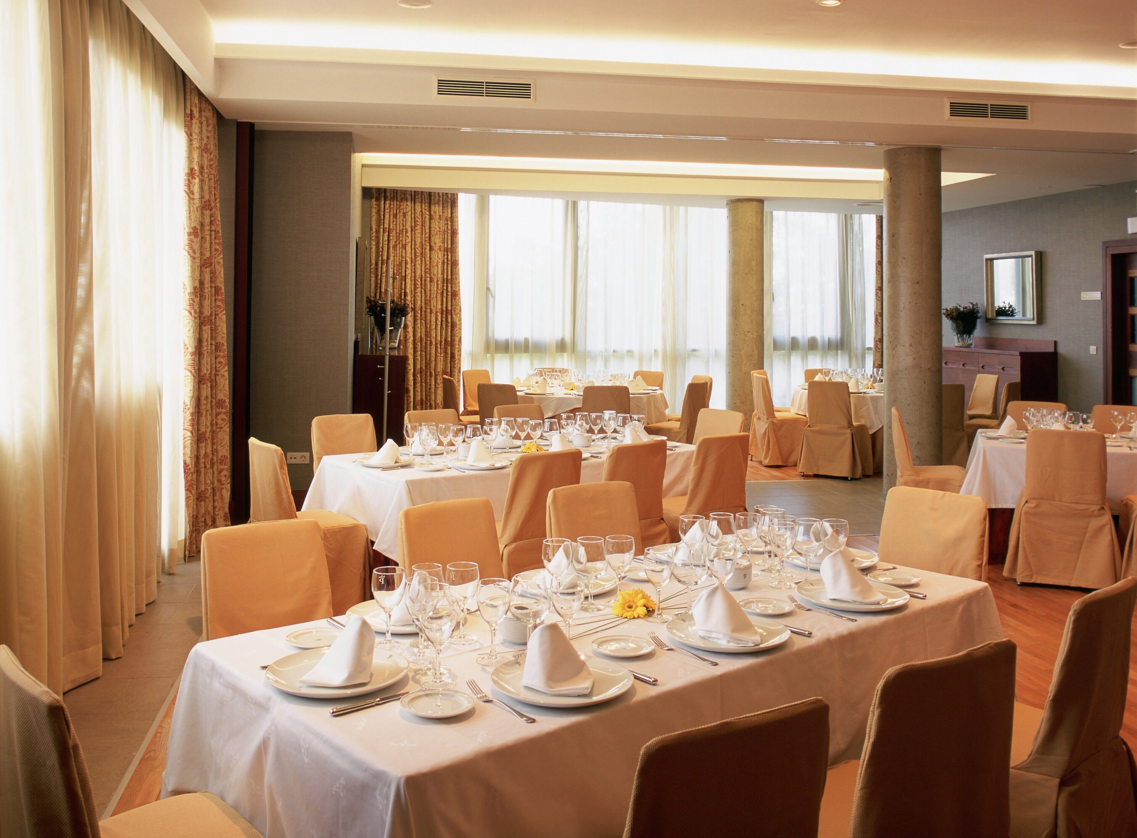 Une salle à manger avec plusieurs tables dressées pour un événement formel, avec des nappes blanches, de la vaisselle, des verres et des chaises disposés avec soin.