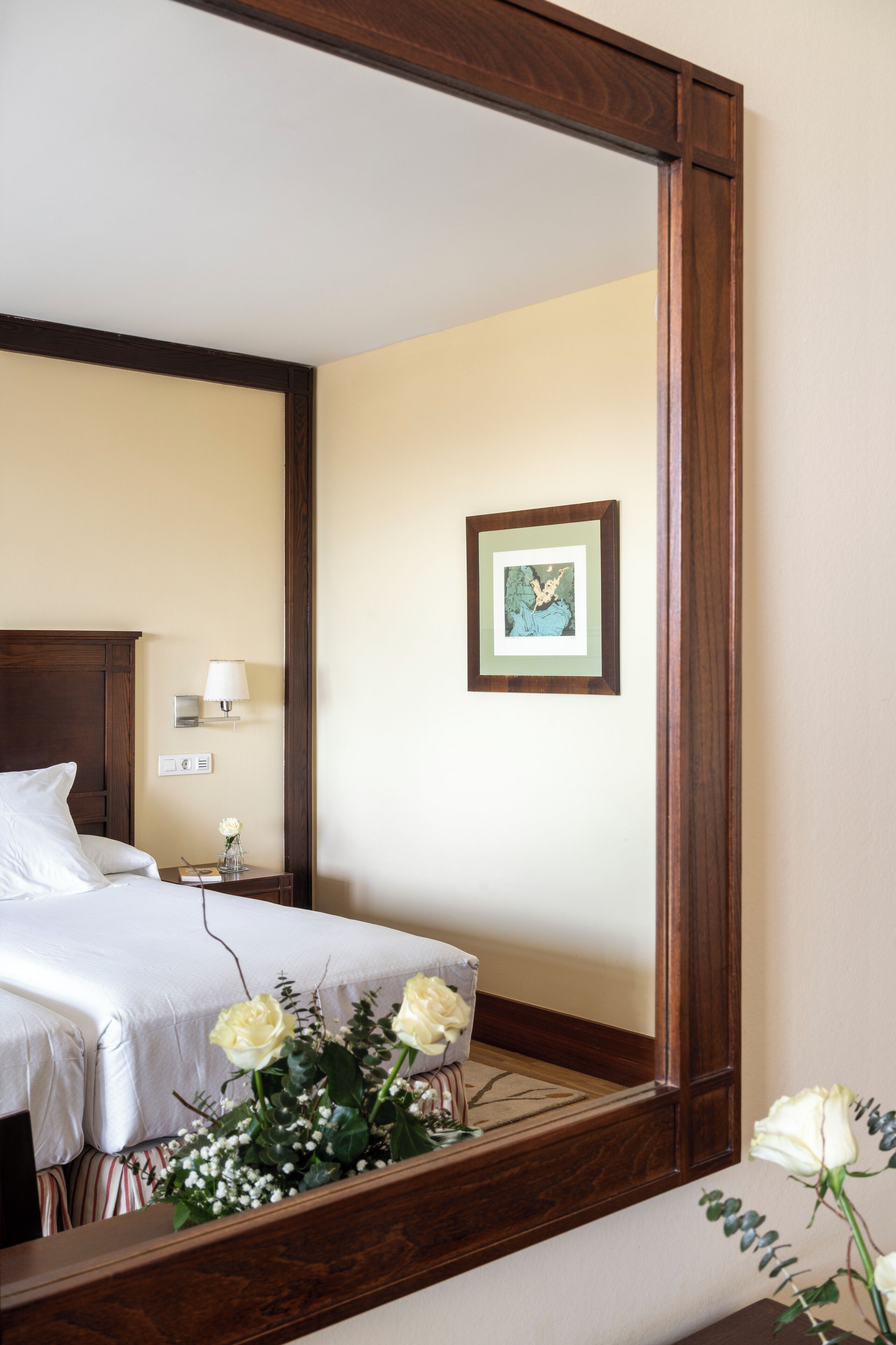 Une chambre d’hôtel avec une tête de lit en bois, un lit recouvert de draps blancs, une table de chevet avec une lampe, un tableau encadré au mur et un vase rempli de fleurs posé sur le sol.