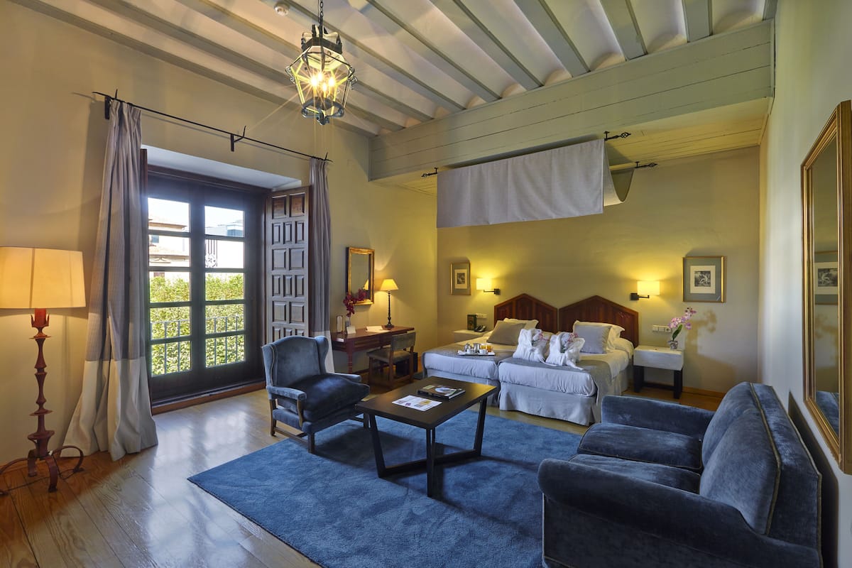 A hotel room with a wooden ceiling, a large window with curtains, a sofa, a chair, a small table, and a bed with a headboard.