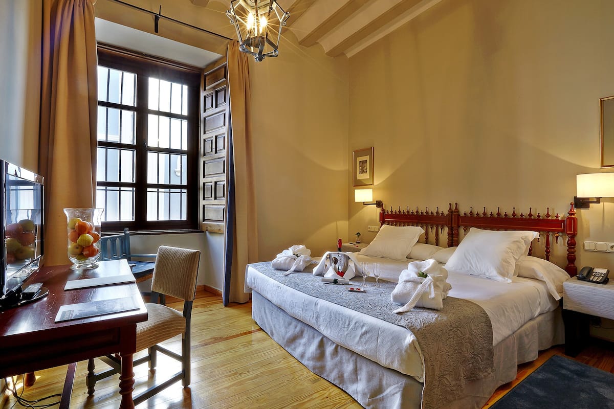 A hotel room with a large bed, a desk with a vase of fruit, a chair, and a window with curtains.