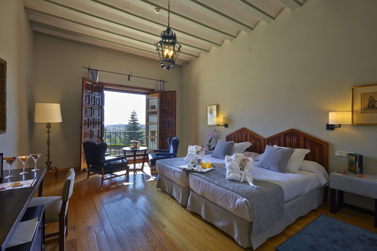 A hotel room with a bed, a desk, a chair, a dining table with a tray of breakfast, and a view of the outdoors.