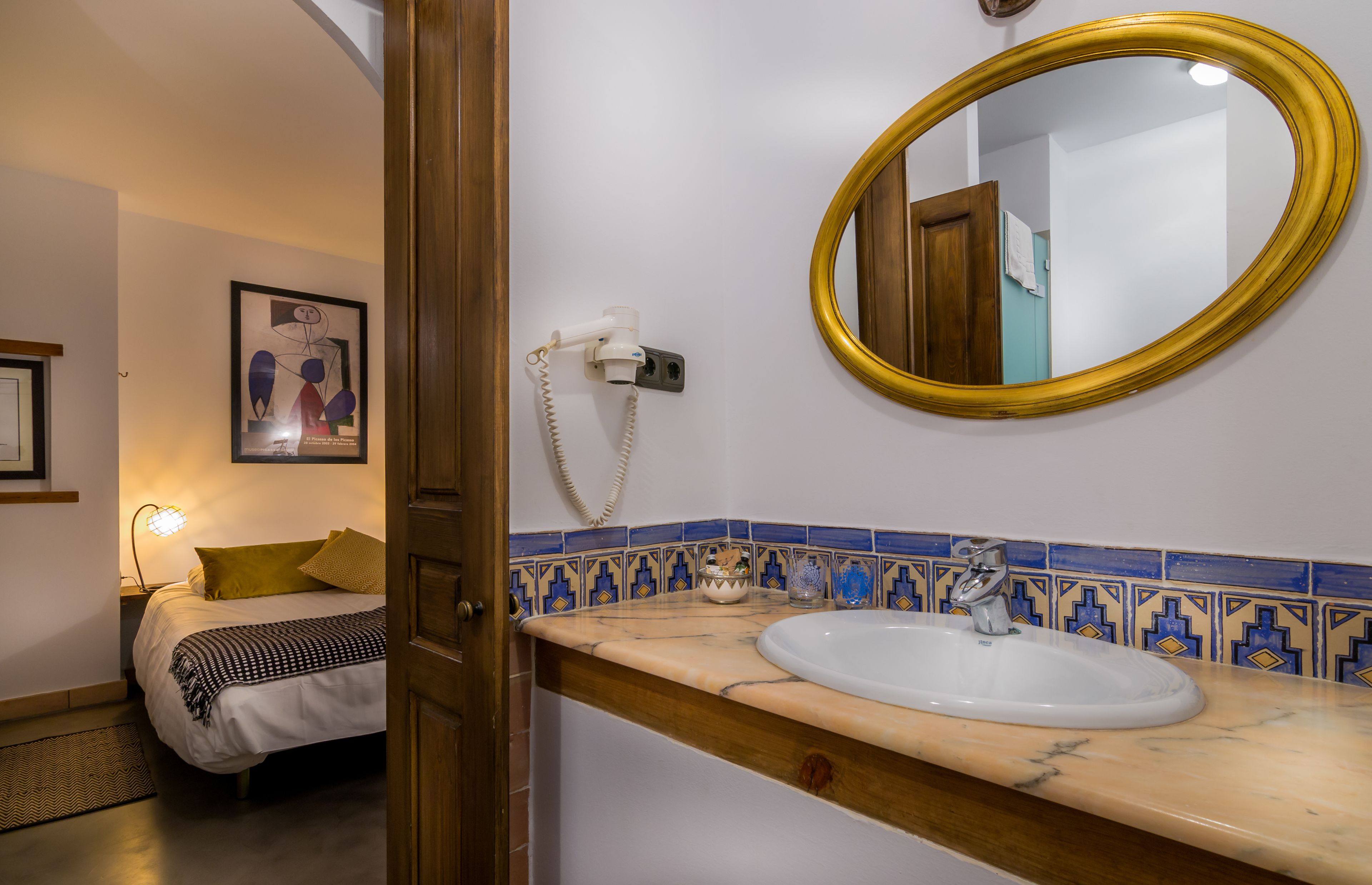 Een hotelbadkamer met een wit wastafelmeubel, een spiegel met gouden lijst en een blauw-geel betegelde achterwand.