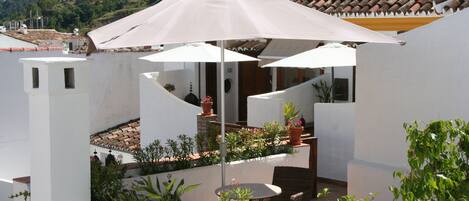 A courtyard with white walls, terracotta roof tiles, and potted plants. There is a round table with chairs and a large umbrella providing shade.