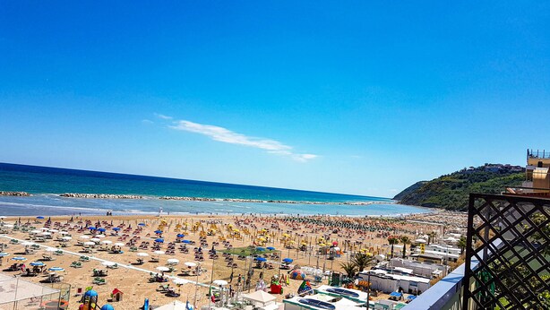 A beach with numerous umbrellas and sunbeds, a clear blue sky, and a distant hill with buildings. - Hotel Cavalluccio Marino (Gabicce Mare)
