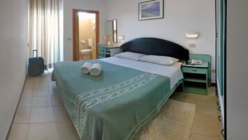 Une chambre d’hôtel avec un lit, une table de chevet, une chaise, une valise et une salle de bain visible à travers une porte ouverte.