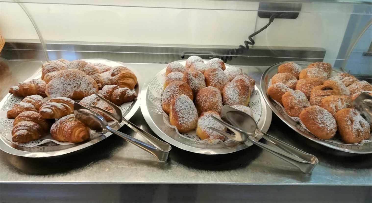 Una teca con tre vassoi di pasticceria, tra cui croissant e altri dolci lievitati, spolverati di zucchero a velo.