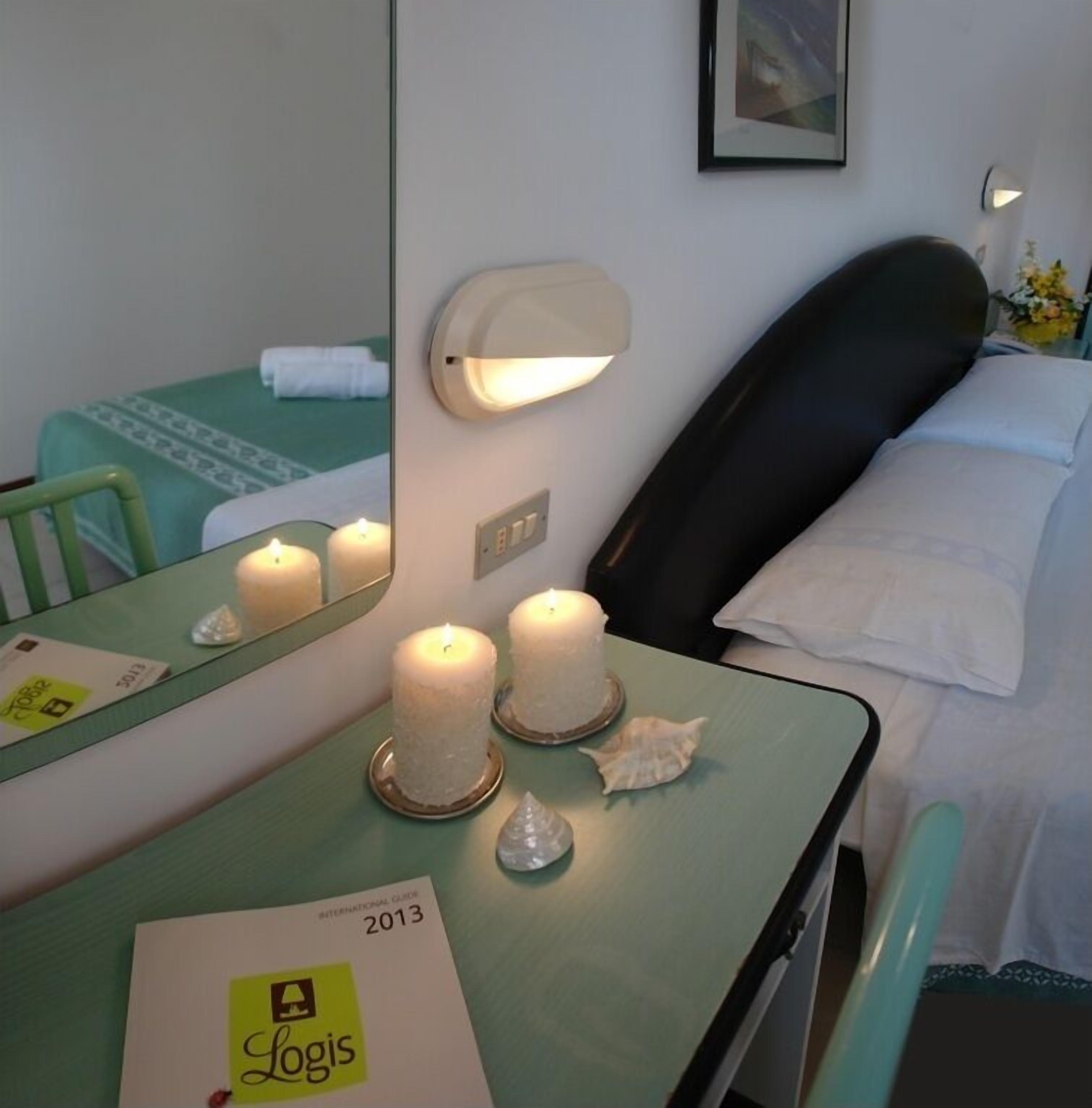 A hotel room with a bed, a bedside table with candles, a mirror, and a small desk with a notebook and a brochure.