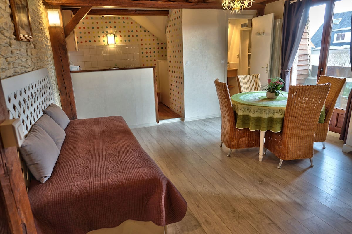 Ein gemütliches Zimmer mit Holzboden, einem Esstisch mit Korbstühlen, einem roten, gepolsterten Sofa und einer Küchenzeile mit farbenfroher Spitze als Rückwand.