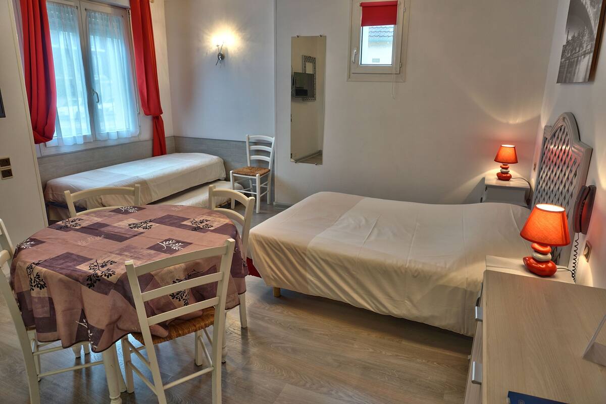 Ein Zimmer mit zwei Einzelbetten, einem Tisch mit Tischdecke, einem Stuhl, einer Lampe, einem Spiegel und einem Fenster mit Vorhängen.