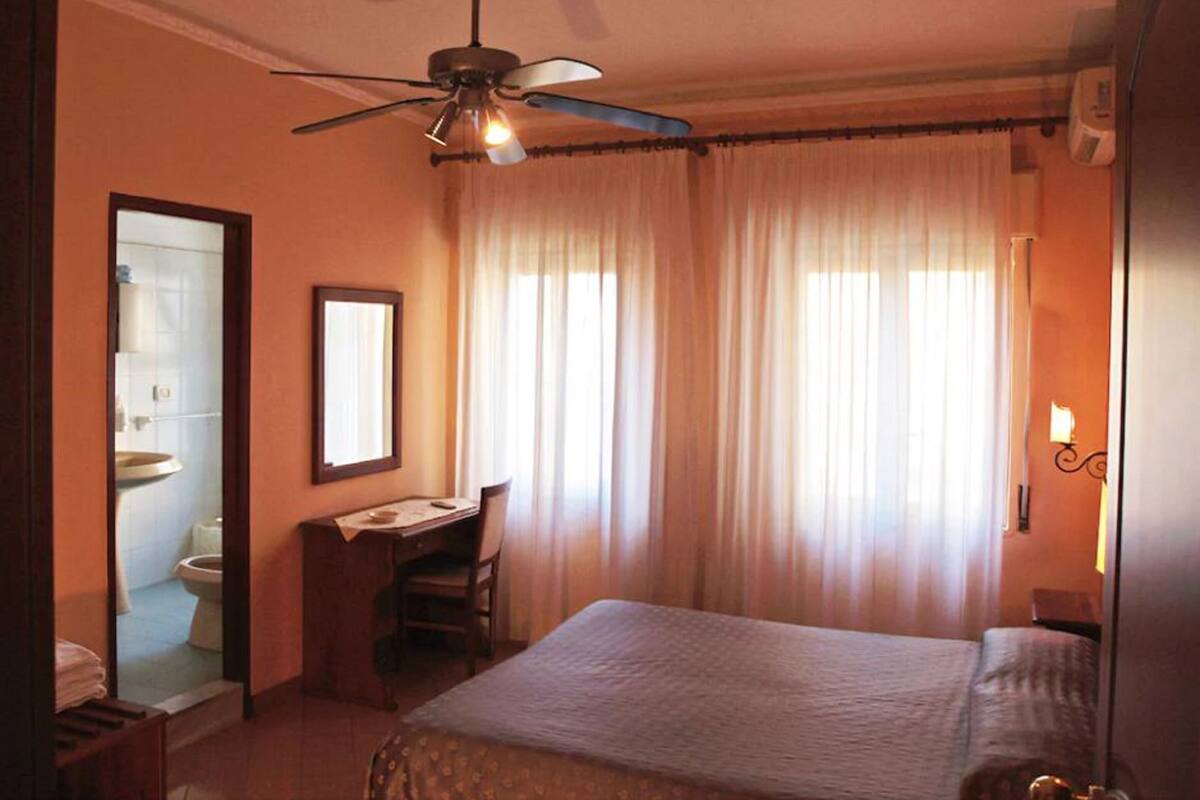 Habitación de hotel con cama, escritorio, silla, ventilador de techo y baño visible a través de una puerta abierta.