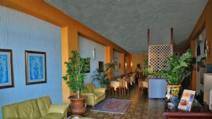 A hotel lobby with a green sofa, a coffee table, a patterned rug, and a reception desk with a plant and a magazine. - Hotel Costa Azzurra (Giardini Naxos)
