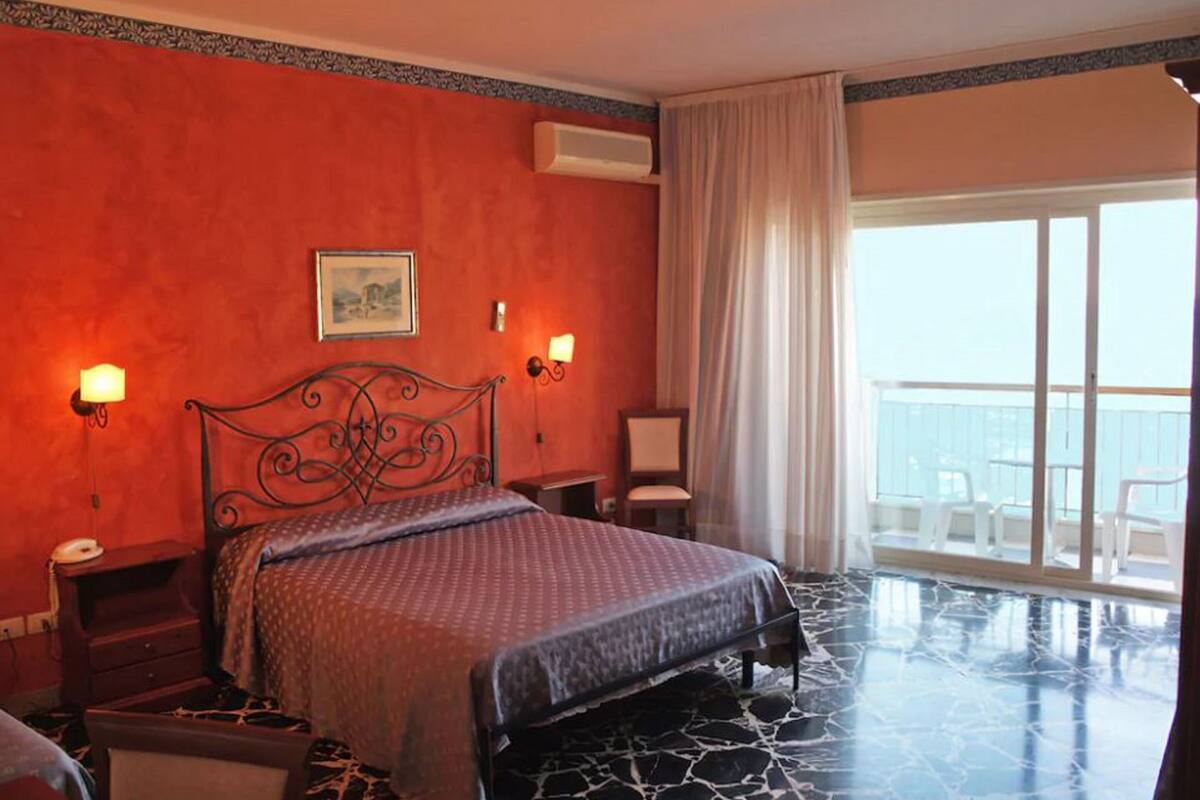 Habitación de hotel con cama, mesitas de noche, una silla, ventana con cortinas y balcón con sillas.