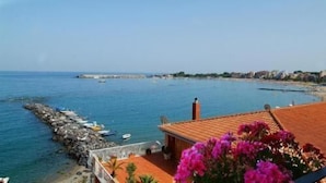A coastal view with a rocky shoreline, boats, and a building with a red roof. - Hotel Costa Azzurra (Giardini Naxos)
