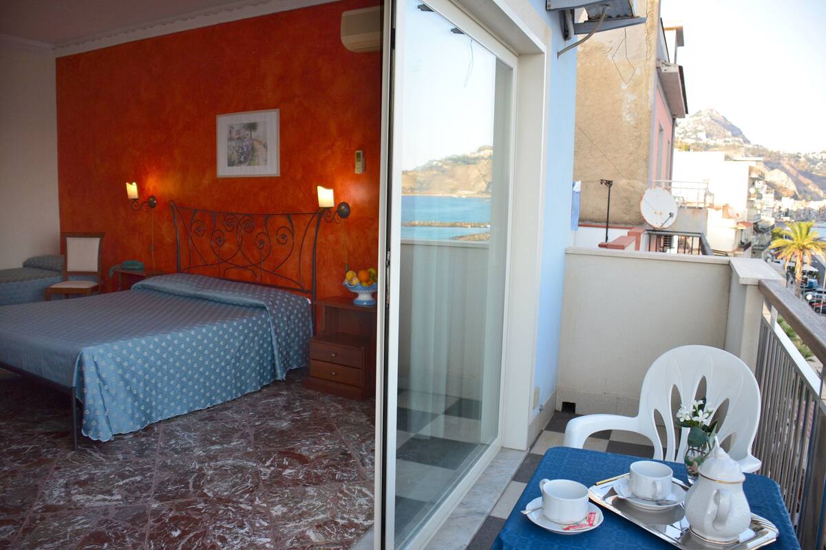 Una habitación de hotel con cama, una mesita con una bandeja de alimentos para el desayuno y un balcón con vistas al mar y a las montañas.