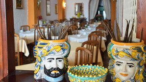 Two ceramic vases with handles, each containing multiple reed sticks, placed on a table in a dining area with wooden chairs and tables. - Hotel Costa Azzurra (Giardini Naxos)