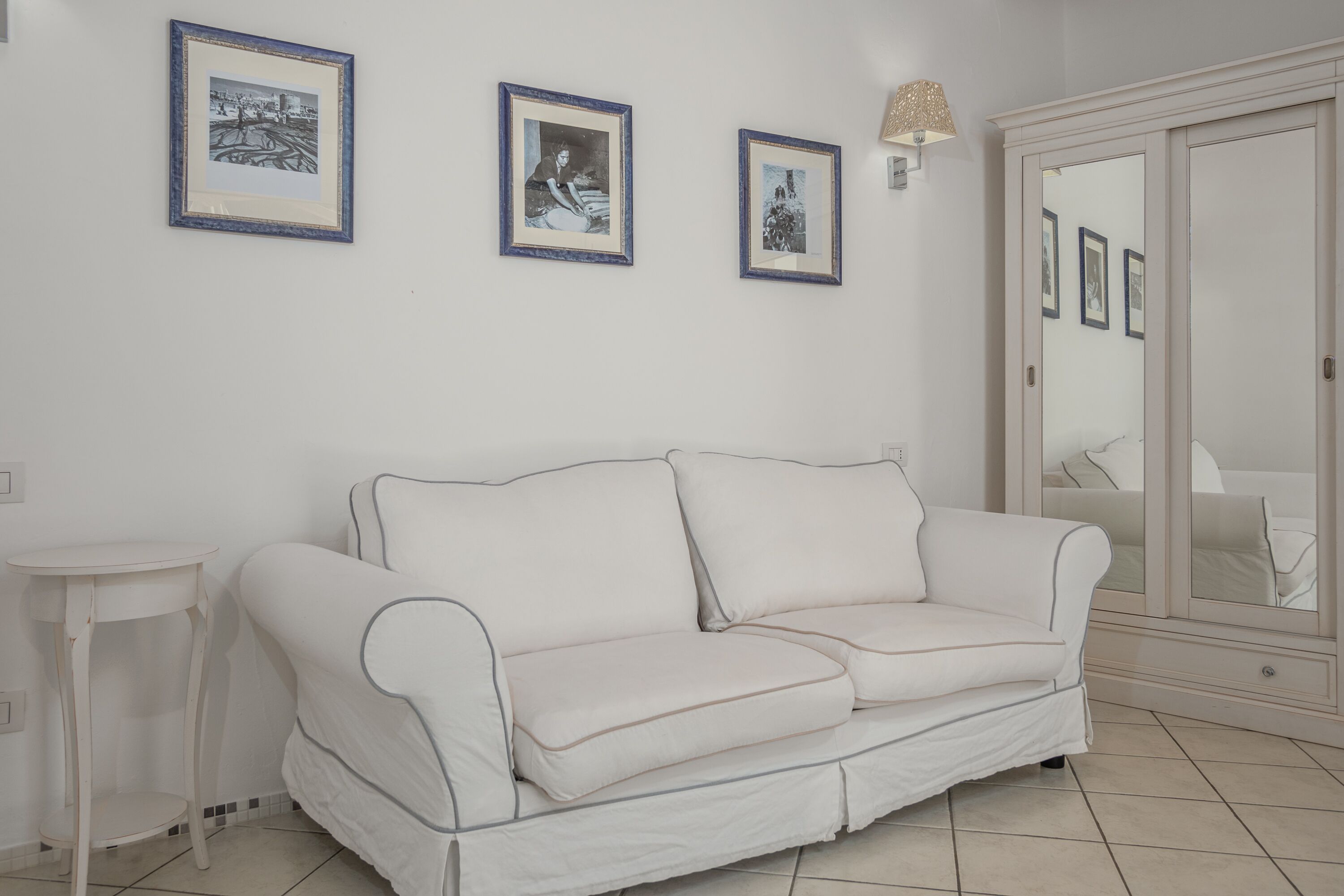 Um sofá branco com uma pequena mesa ao lado, uma parede com quadros e um guarda-roupa com espelho.