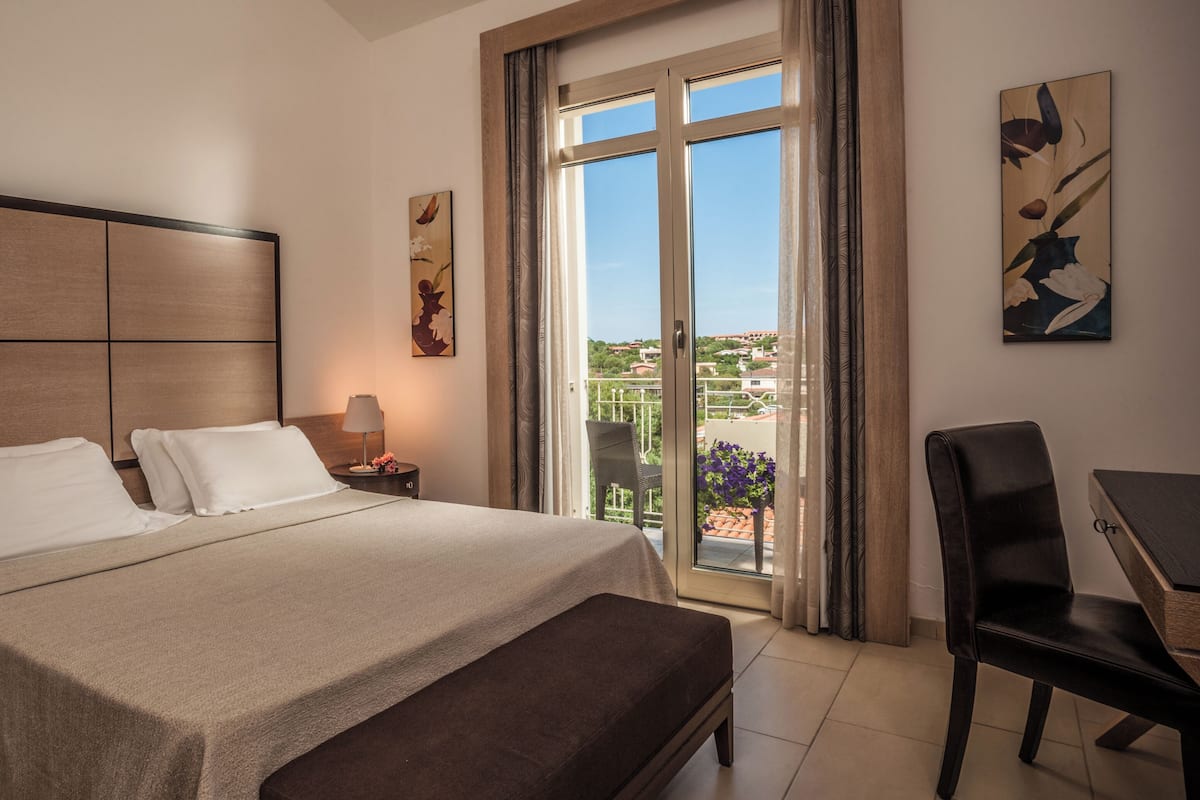 Habitación de hotel con cama, escritorio, silla, mesita de noche y ventana con vistas.
