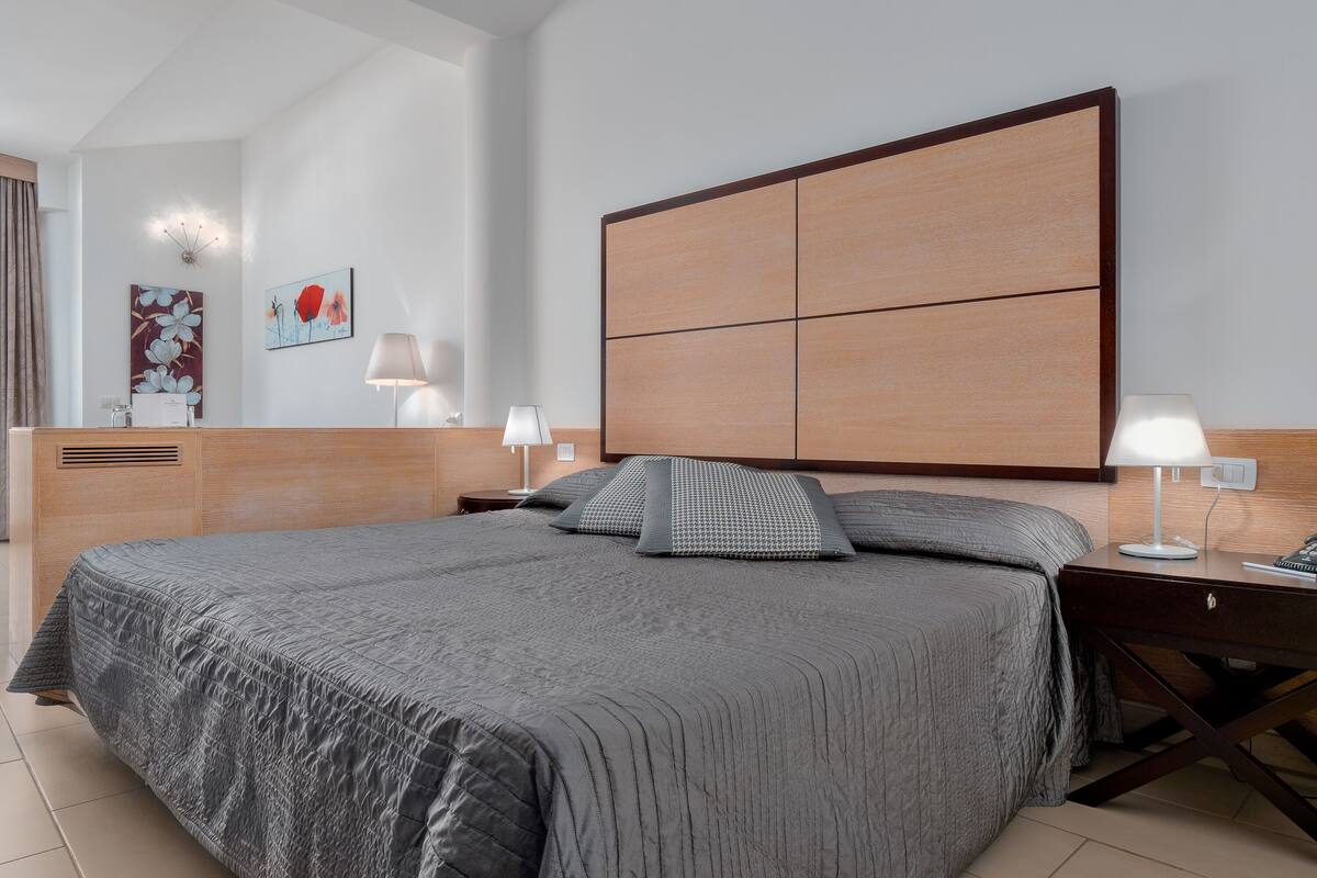 Una habitación de hotel moderna con una cama grande, cabecera de madera, mesitas de noche con lámparas y obras de arte en las paredes.