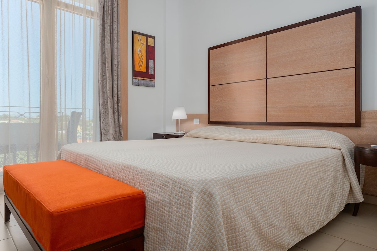 Un dormitorio ordenado con una cama grande, una mesita de noche con lámpara y una ventana con cortinas.