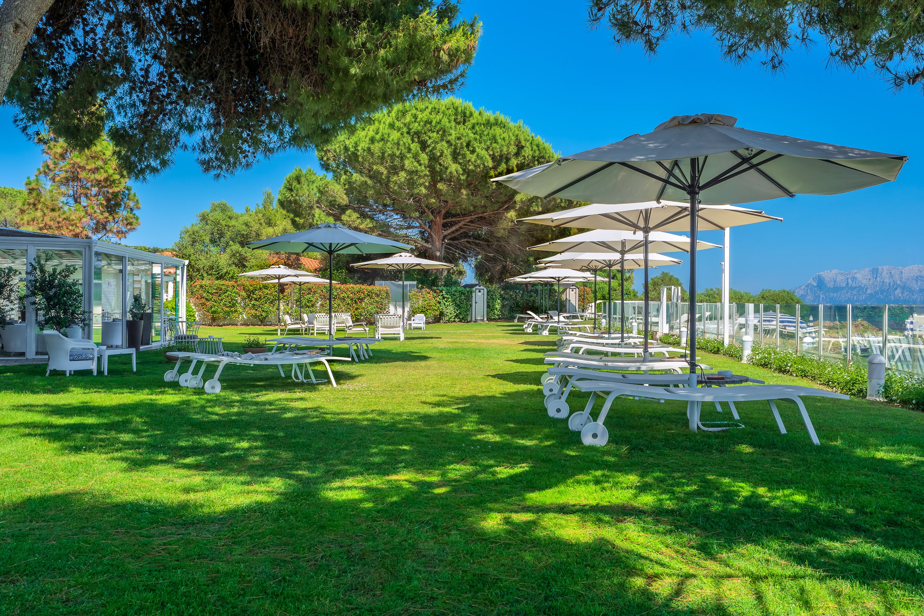 A well-maintained outdoor area with white lounge chairs and umbrellas, a glass-enclosed structure, and a view of the sea.
