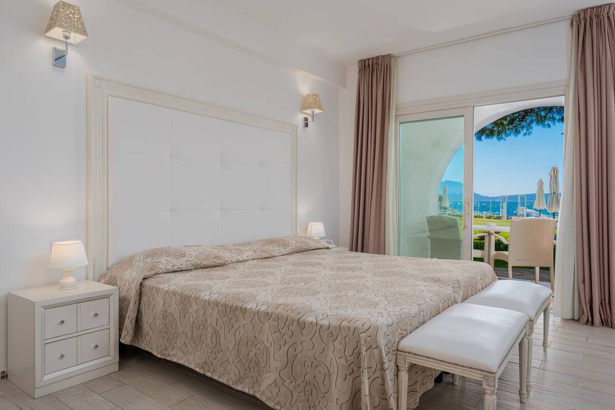 Un dormitorio con una cama grande, una mesita de noche con cajones, un banco y vistas al exterior a través de una puerta de cristal.