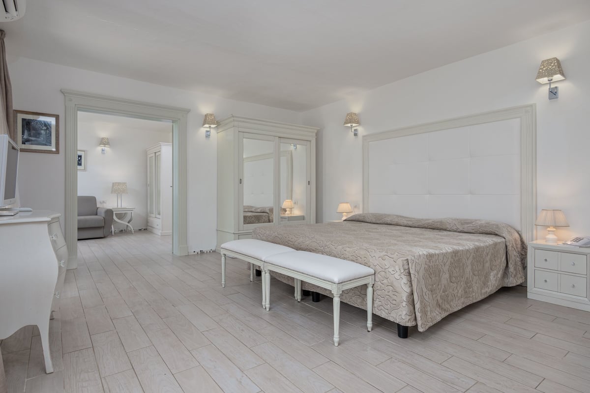 Un dormitorio con una cama grande, un banco, un escritorio y un aparador. Hay dos mesitas de noche con lámparas y un espejo sobre la cama.
