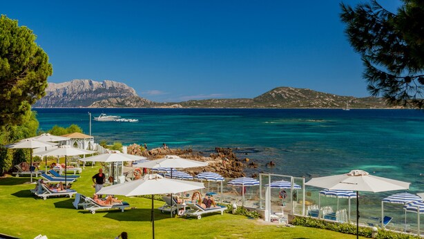 A coastal resort with sun loungers and umbrellas, a clear view of the sea, and rocky hills in the background. - The Pelican Beach Resort & SPA - Adults Only (Olbia)
