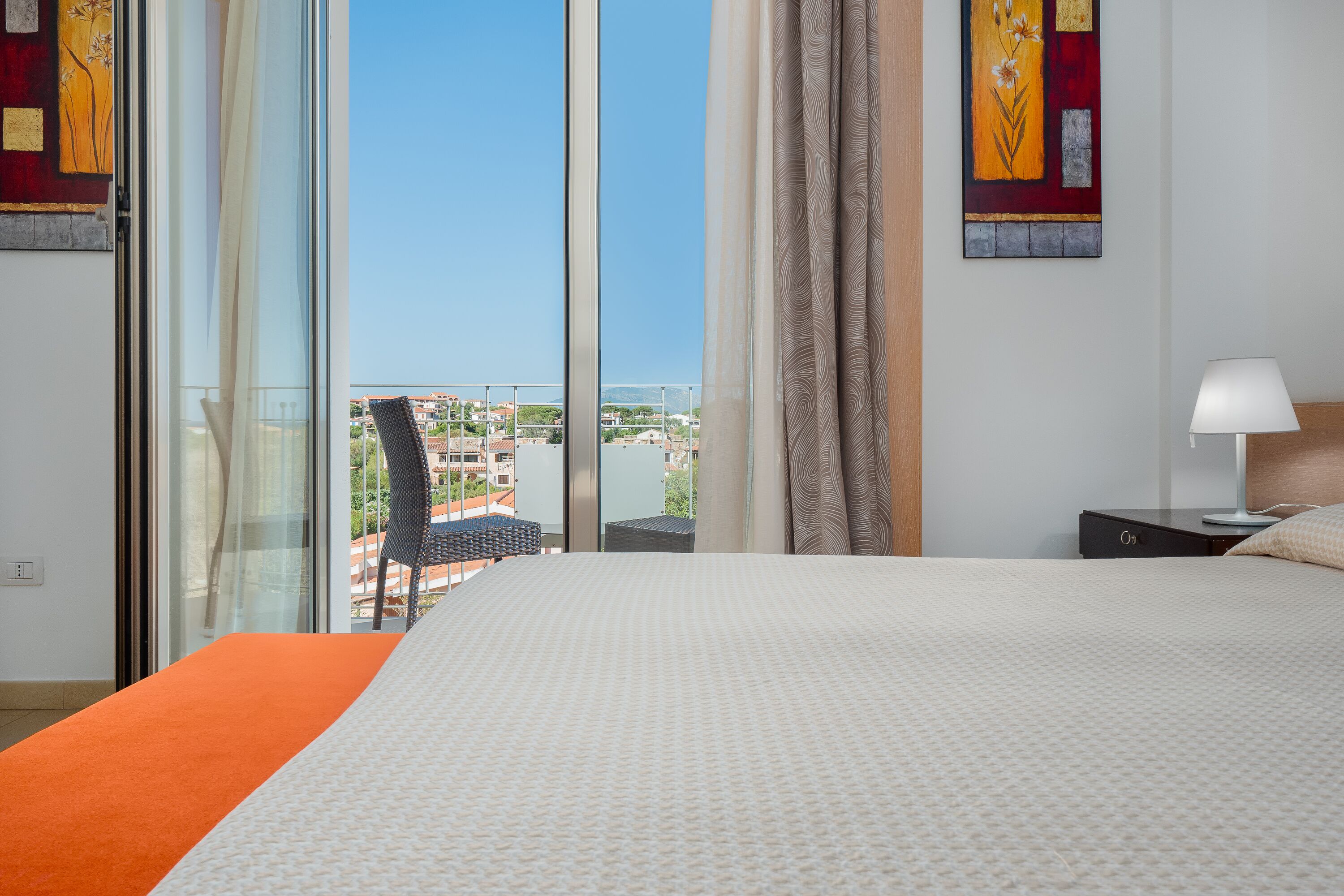 A hotel room with a large bed, a view of the city through floor-to-ceiling windows, a chair, and a nightstand with a lamp.
