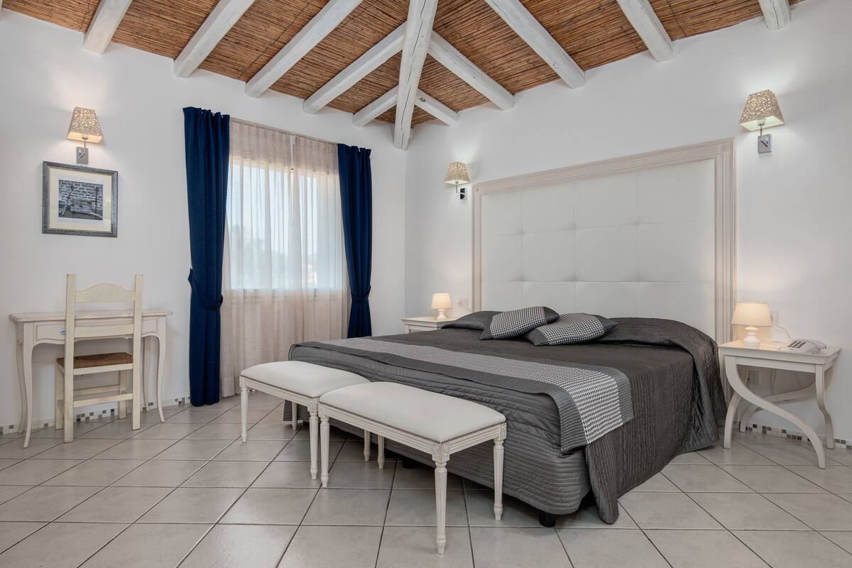 Un dormitorio con cama, banco, mesitas de noche y techo de vigas de madera.