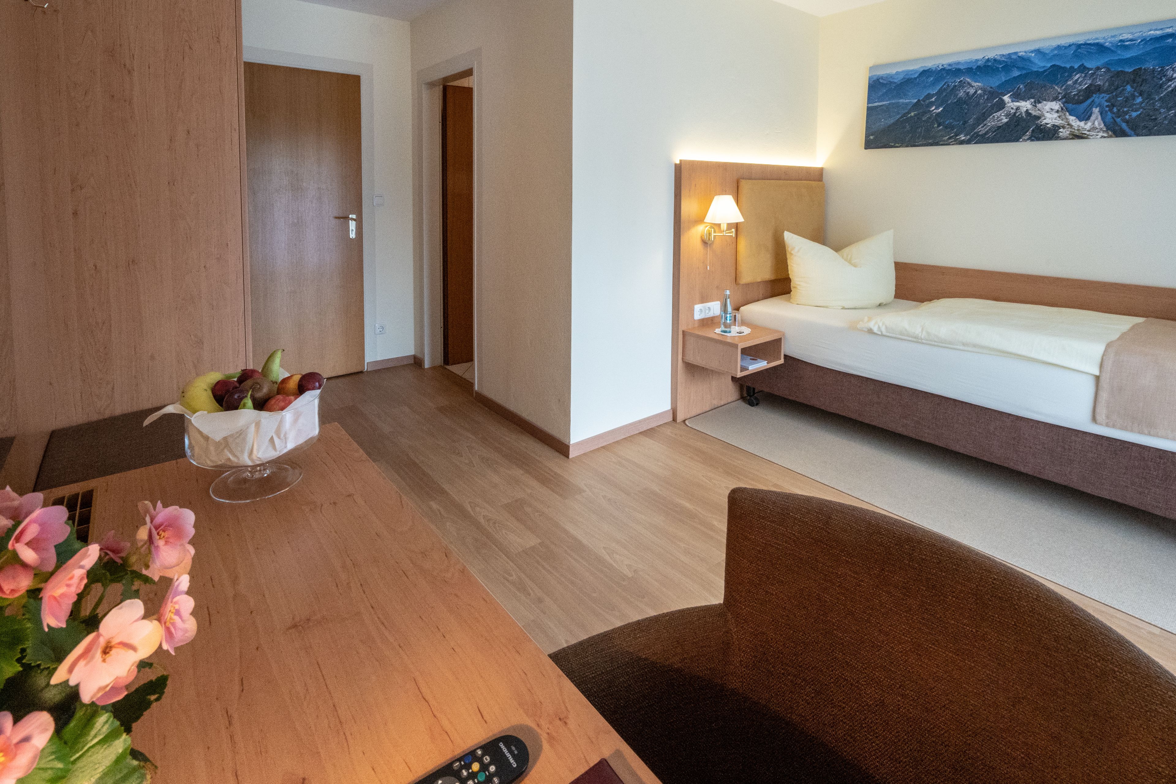 A hotel room with a wooden floor, a bed with a headboard, a bedside table with a lamp, a small table with fruit and flowers, and a painting on the wall.