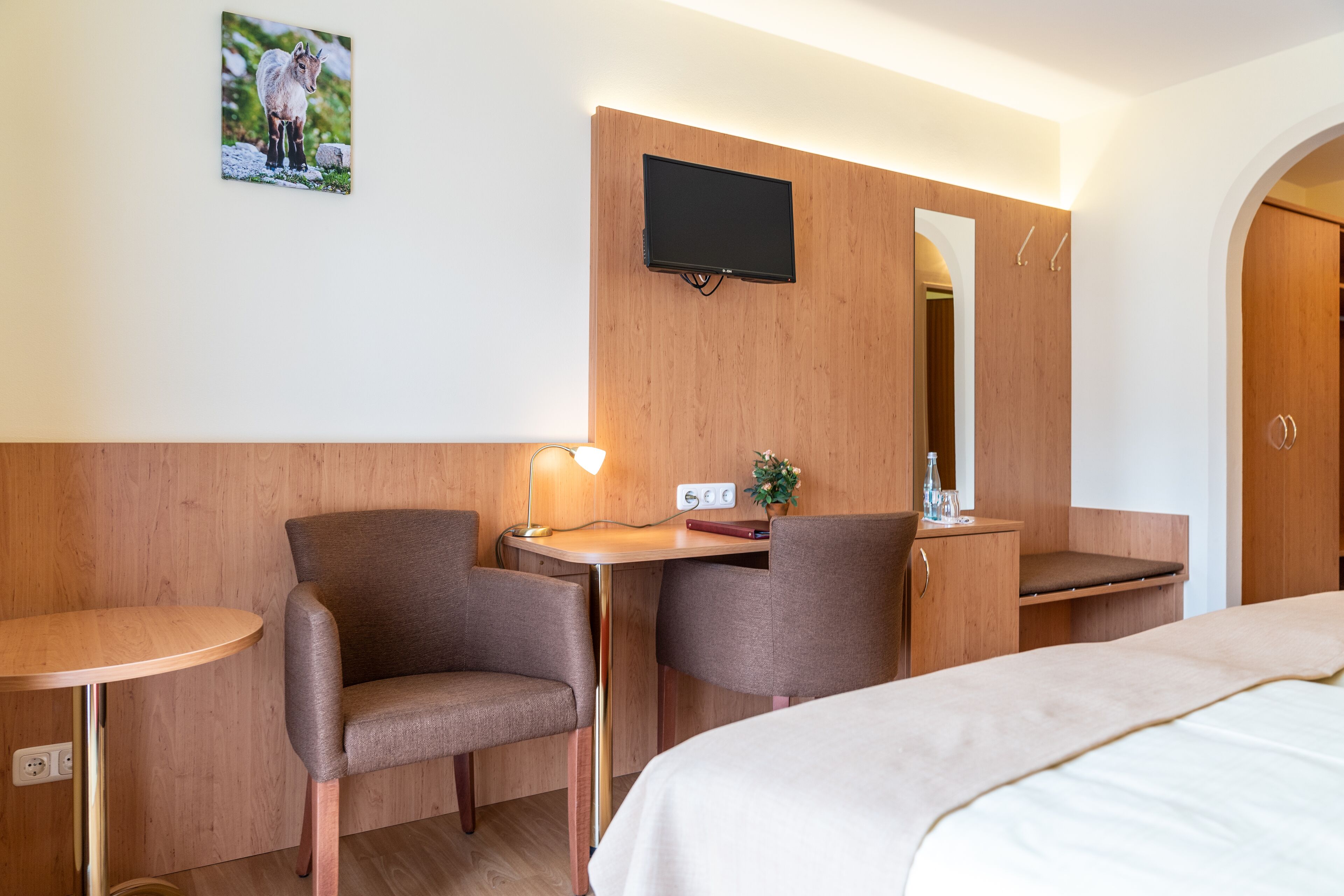 Ein Hotelzimmer mit einem Bett, zwei Sesseln, einem kleinen Schreibtisch mit Lampe, einem an der Wand befestigten Flachbildfernseher und einem Spiegel.