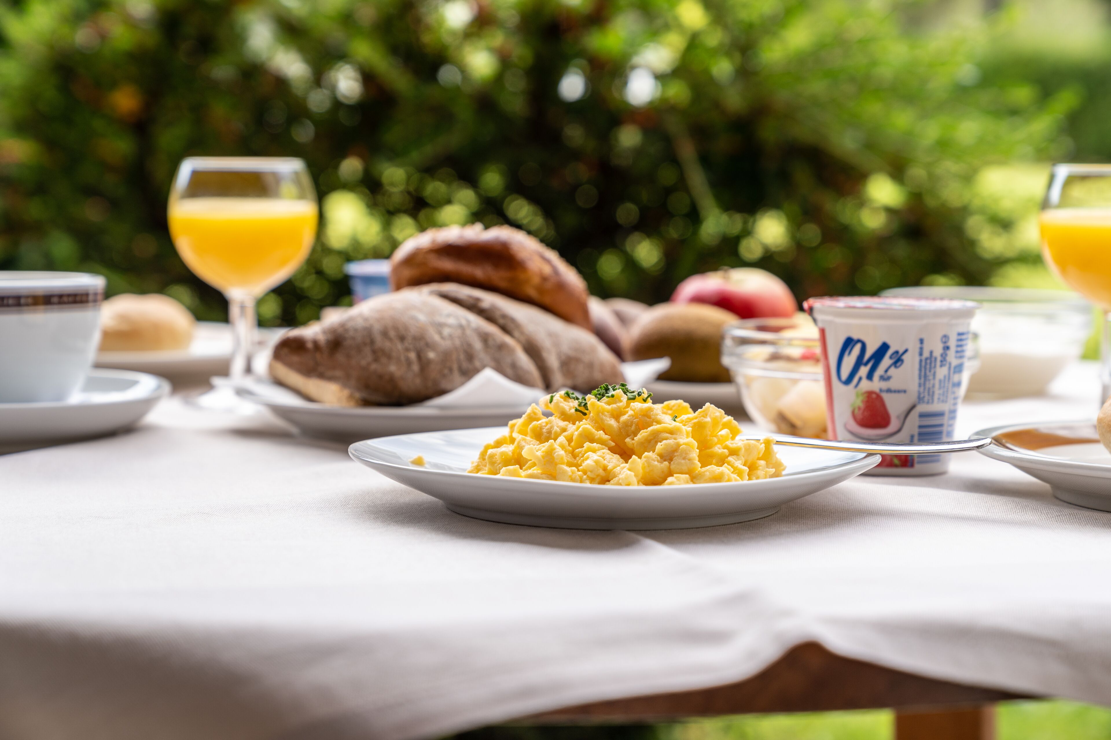 A breakfast table with scrambled eggs, croissants, a glass of orange juice, and a yogurt container.