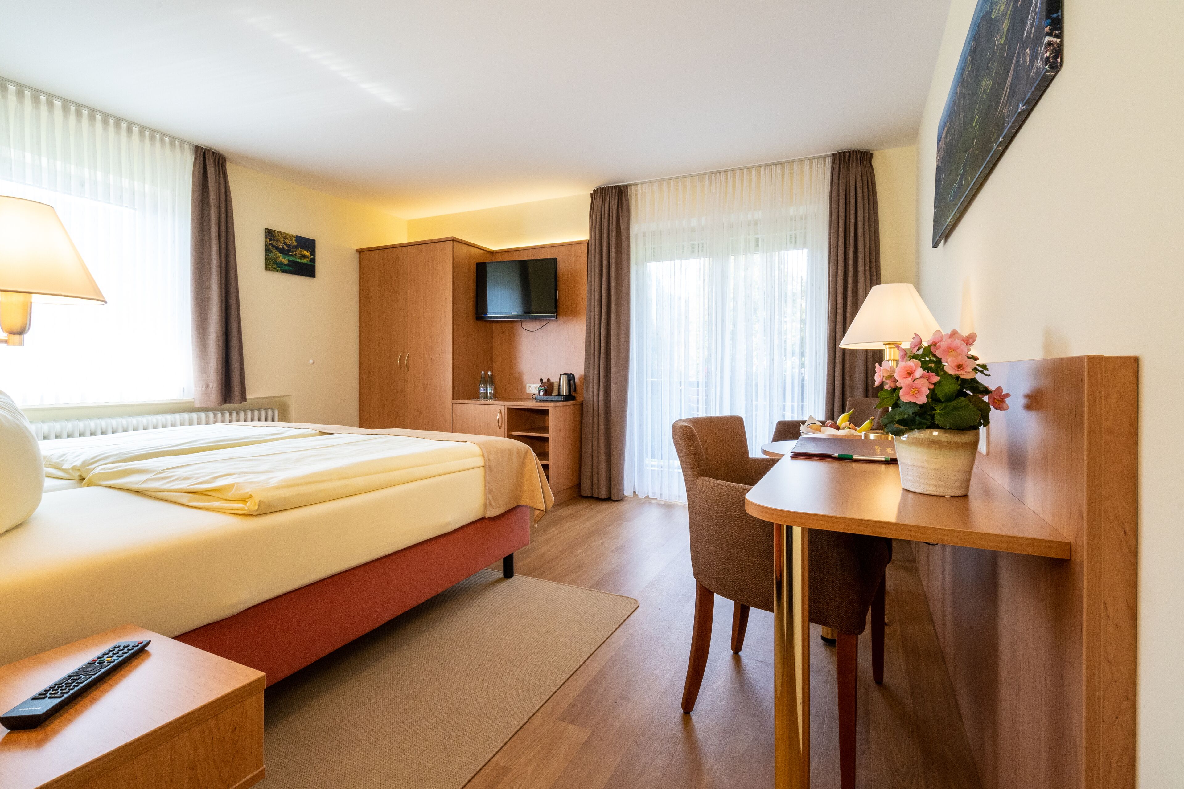 A hotel room with a bed, bedside table, TV, desk with chair, and a small kitchenette area.